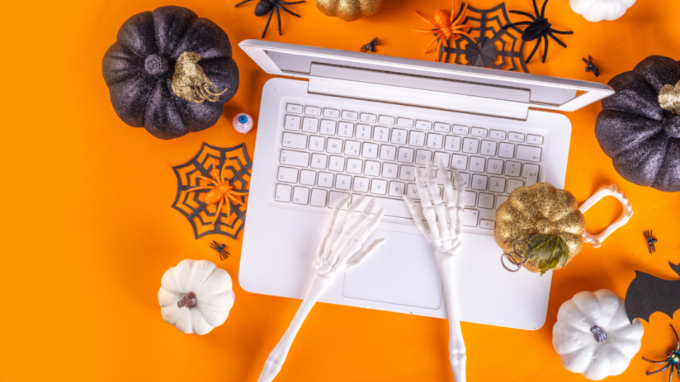 Skeleton hands typing on a white laptop against an orange background, surrounded by black, white, and gold pumpkins, plastic spiders, and spiderwebs. A spooky Halloween vibe merges with the pursuit of job search strategies.